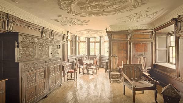 An empty wooden chair beside a low railing in a quiet paneled room with daylight from a high window