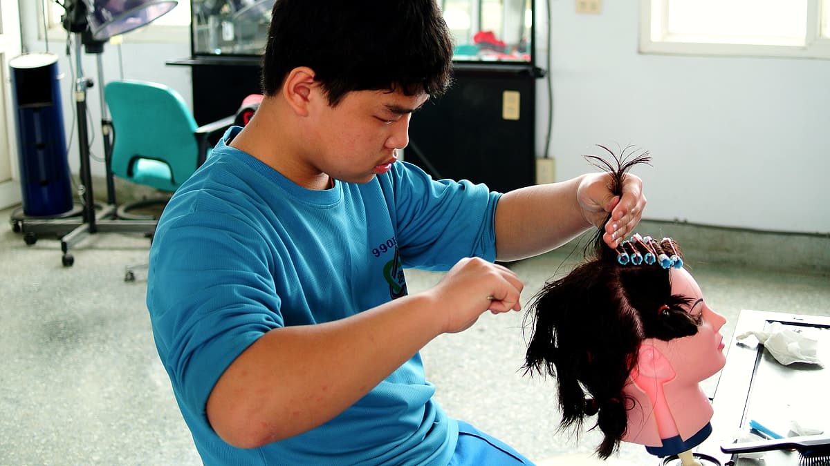 Students at a training salon practicing haircutting on mannequin heads clamped to a row of workstations